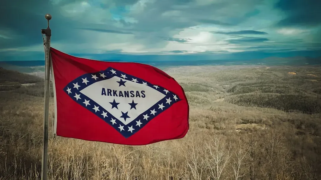 An Arkansas flag flying in the wind with an open field and cloudy skies.