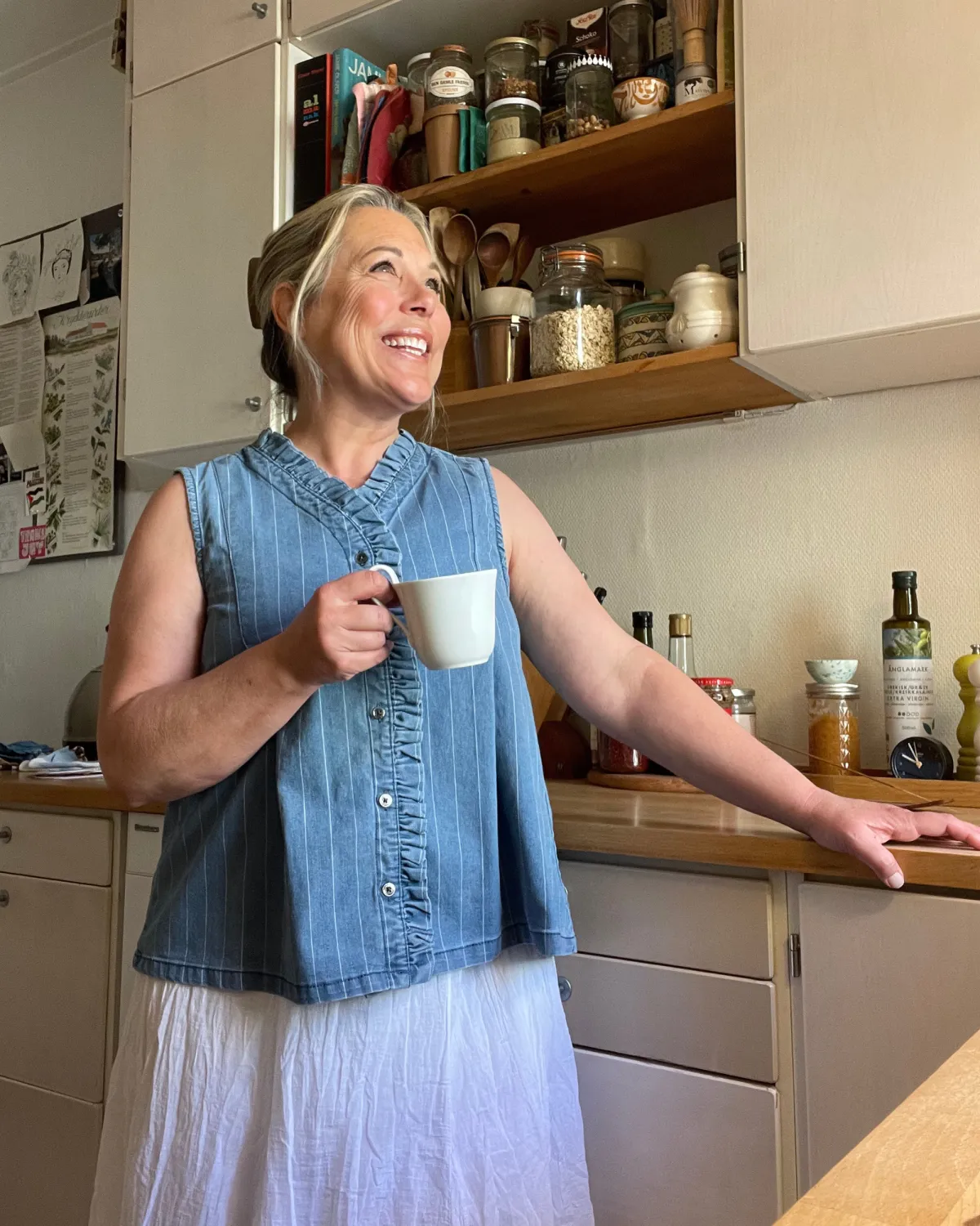 Pasha smiling with a cup of tea, dressed casually in a denim shirt and linen skirt
