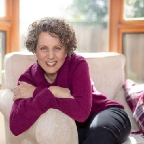Portrait of a smiling woman with curly hair sitting on a light-colored armchair by a window, wearing a deep pink top.