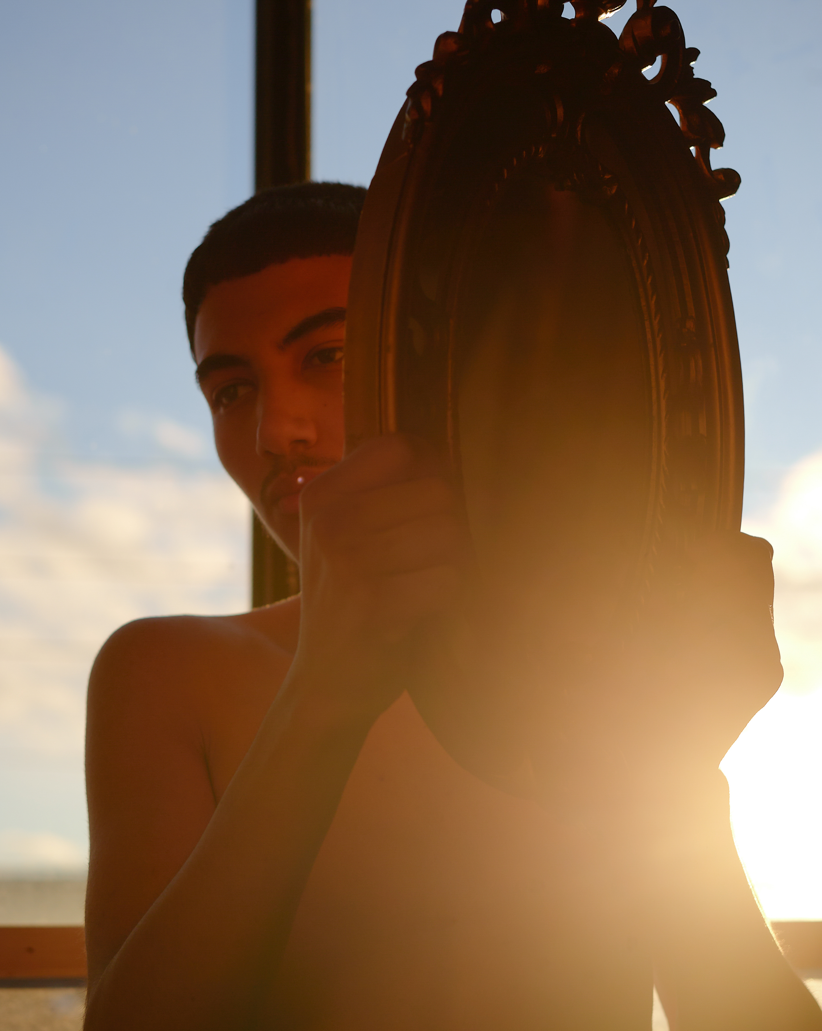 Cinematic portrait of a young man looking into an ornate vintage oval mirror, reflecting his face against a backdrop of clouds and sky at dusk.