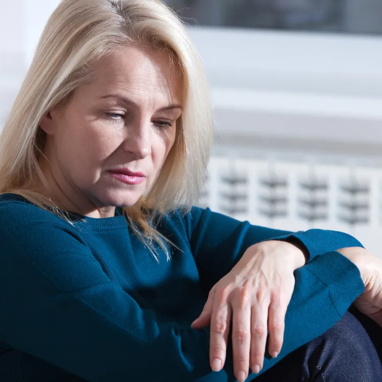 Woman wrapped in a soft blanket, sitting by a window with a cup of coffee, gazing thoughtfully outside, representing quiet reflection and gentle support for women navigating grief.