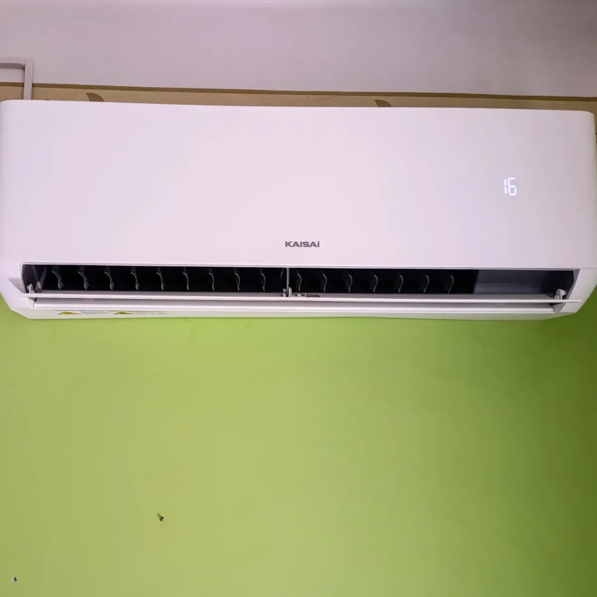 Egy KAISAI feliratú, beltéri klímaberendezés egy világoszöld falon. A kijelző 16-os hőmérsékletet mutat.