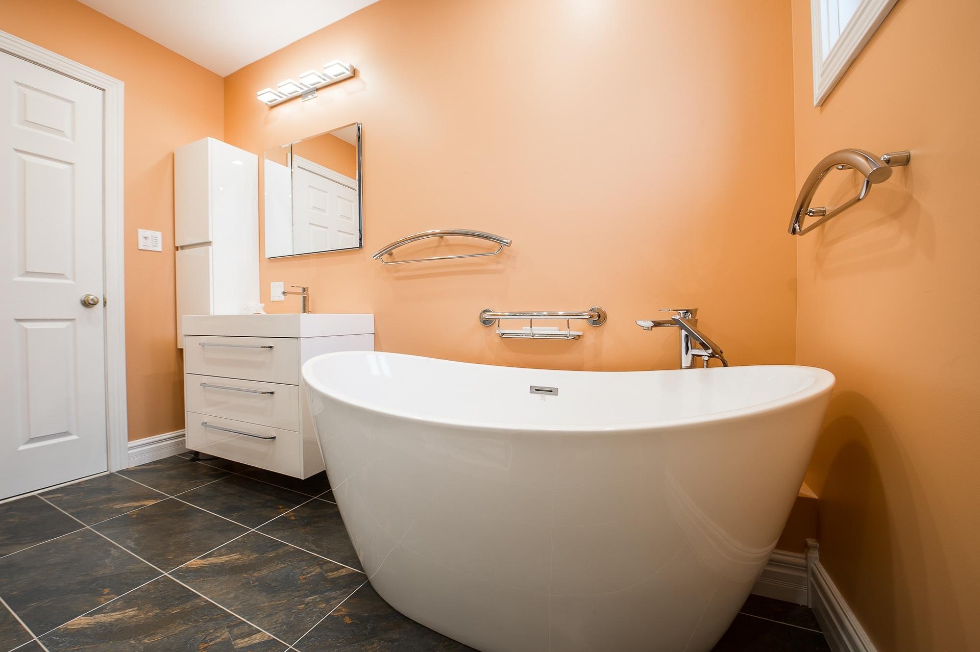 orange walled bathroom with white bathtub, vanity, cupboard and door