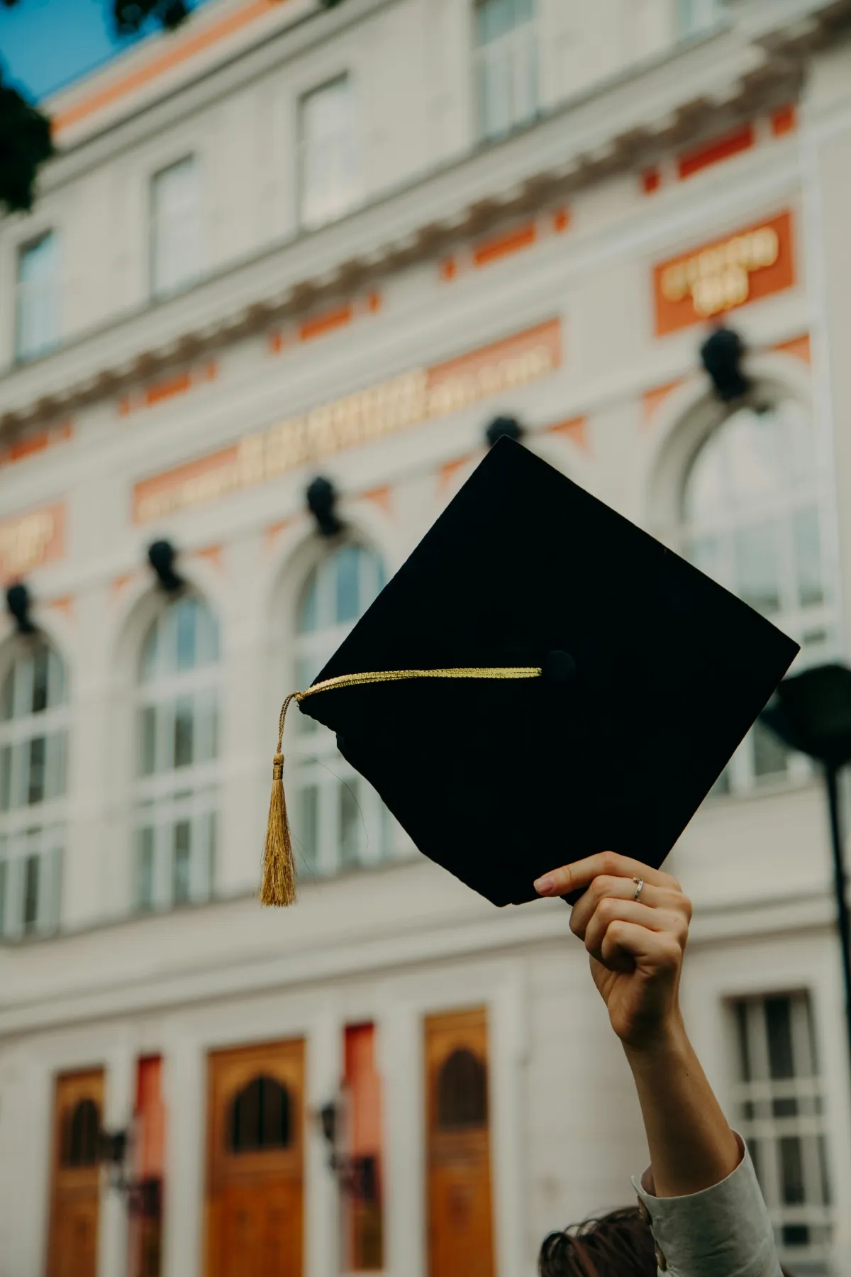 Hand holding a rolled certificate tied with a red ribbon next to a graduation cap
