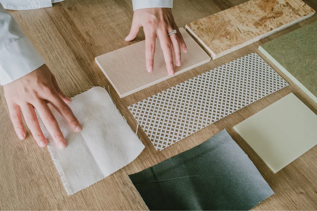 Interior designer's hand selecting sustainable materials including bamboo wood, recycled wood panels, cork tiles, and various eco-friendly fabrics on a wooden table. The collection is arranged to highlight texture and color variety, emphasizing eco-conscious choices in interior design.