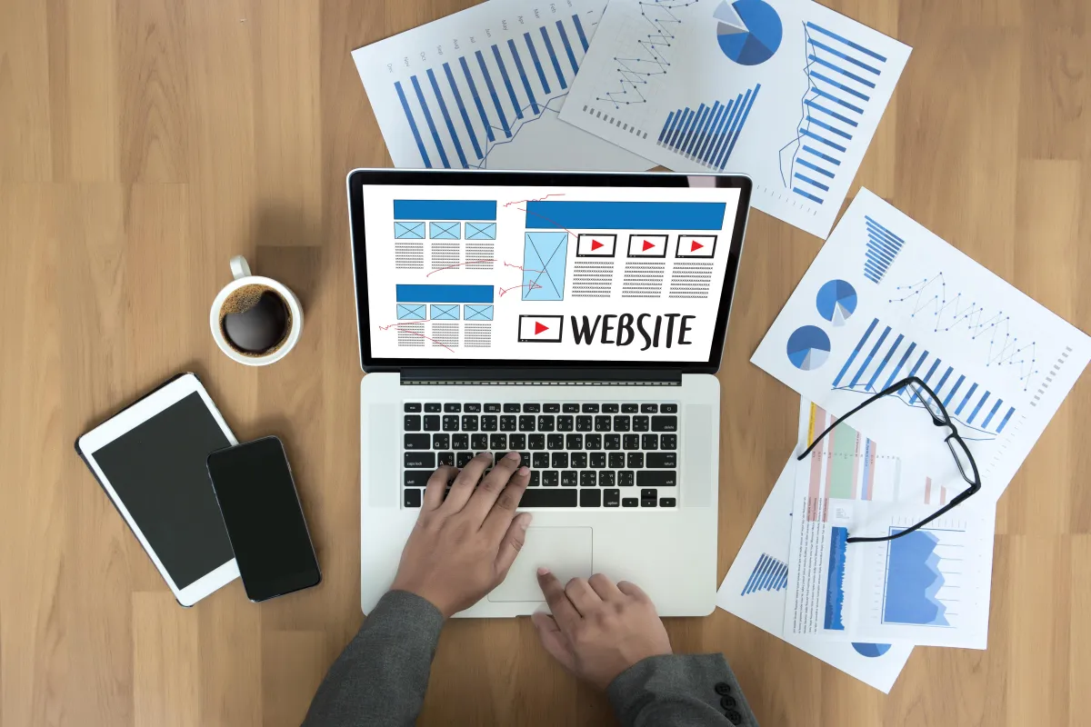Top-down view of a desk with a person working on a laptop showing a website wireframe, surrounded by charts, graphs, tablet, smartphone, coffee, and eyeglasses, representing web development and data-driven design.