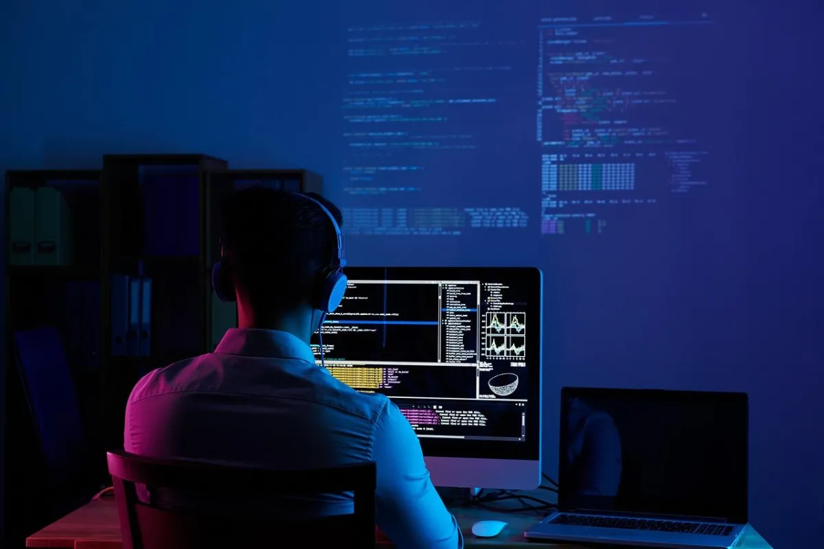 Back view of a developer wearing headphones and working on multiple screens with code and data visualizations in a dark room, representing software development and cybersecurity.