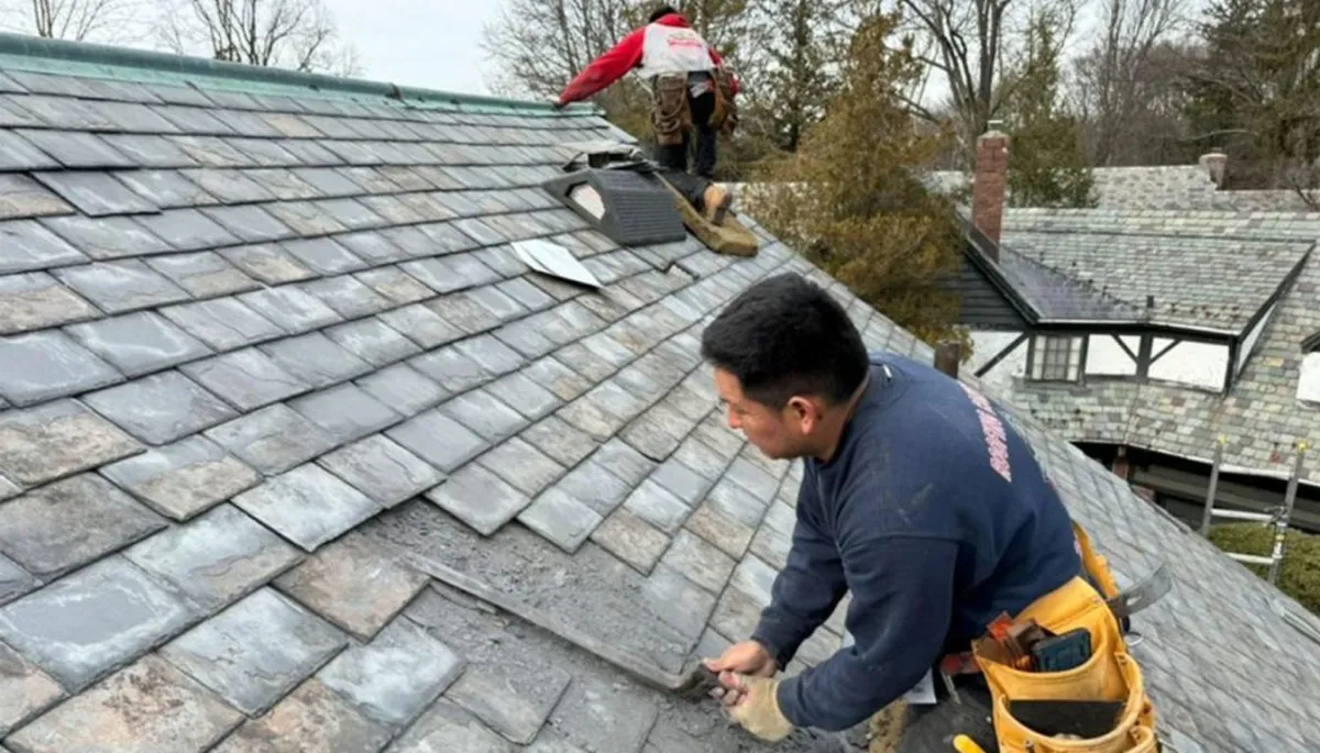 Two men installing slate tiles on a residential roof. One man is in the foreground applying tiles, while the other works further up the roof. 