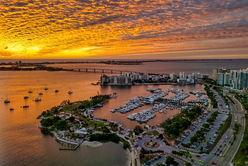 Aerial view of Sarasota, Florida waterfront at sunset, featuring a marina filled with boats, coastal skyline, bridge, and glowing orange clouds over the bay.