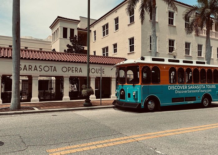 Street view of the Sarasota Opera House in downtown Sarasota, Florida, with a colorful sightseeing trolley parked in front of the historic building lined with palm trees.