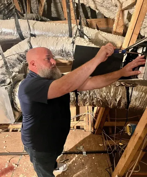 A man is installing ductwork in an attic, surrounded by insulation and beams.
