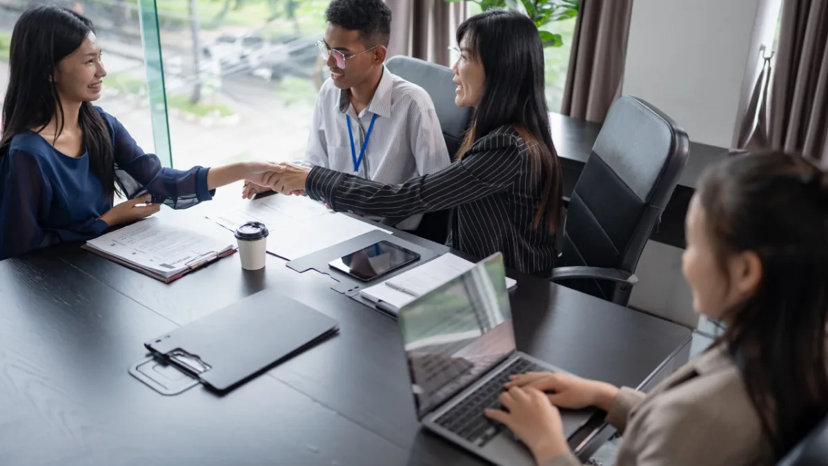 Three business professionals shaking hands across conference table while fourth person works on laptop, with documents and coffee cup on table in bright modern office, representing successful talent attraction, cultural fit hiring, and positive workplace culture that retains top employees.