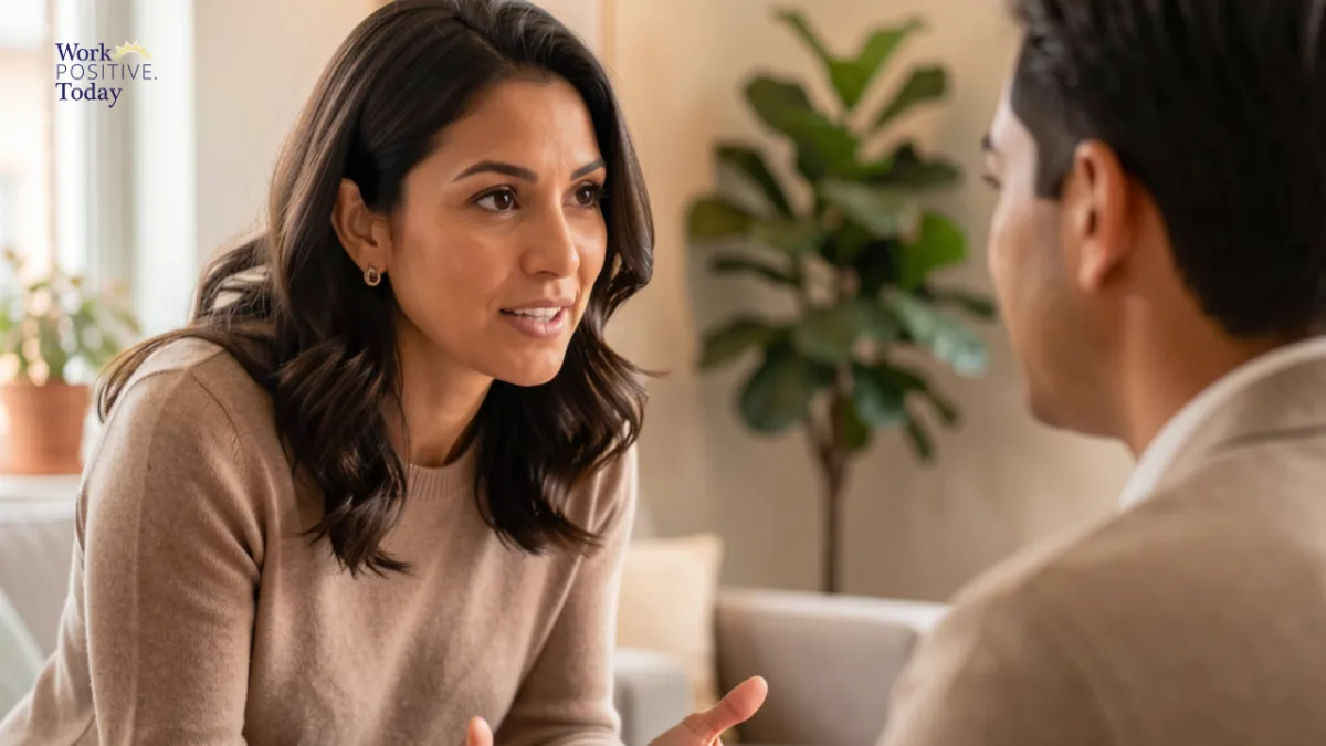Professional woman in beige sweater engaged in empathetic one-on-one conversation with colleague in warm, bright office setting with plants, demonstrating active listening with open body language and genuine eye contact, illustrating leadership empathy that helps employees picture staying 2.5 years longer, with Work Positive Today logo in corner.