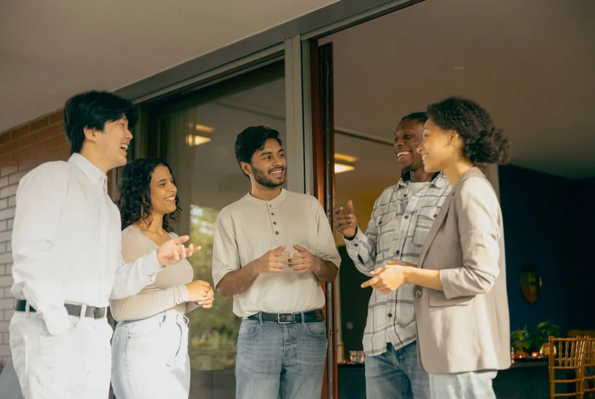 Majority-Hispanic group of college students networking and socializing indoors — mixed genders and ethnicities in casual-professional attire, warm indoor lighting and clear indoor cues (walls and lamps), upbeat and engaged atmosphere