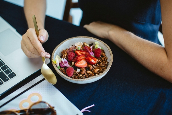 a woman sits at a computer (face unseen) with a bowl of fruit topped food