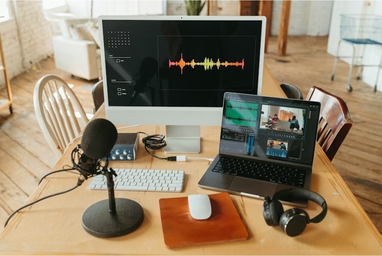 A wooden desk filled with different electronics, including a monitor, keyboard, mic, mixer, laptop, and a mouse