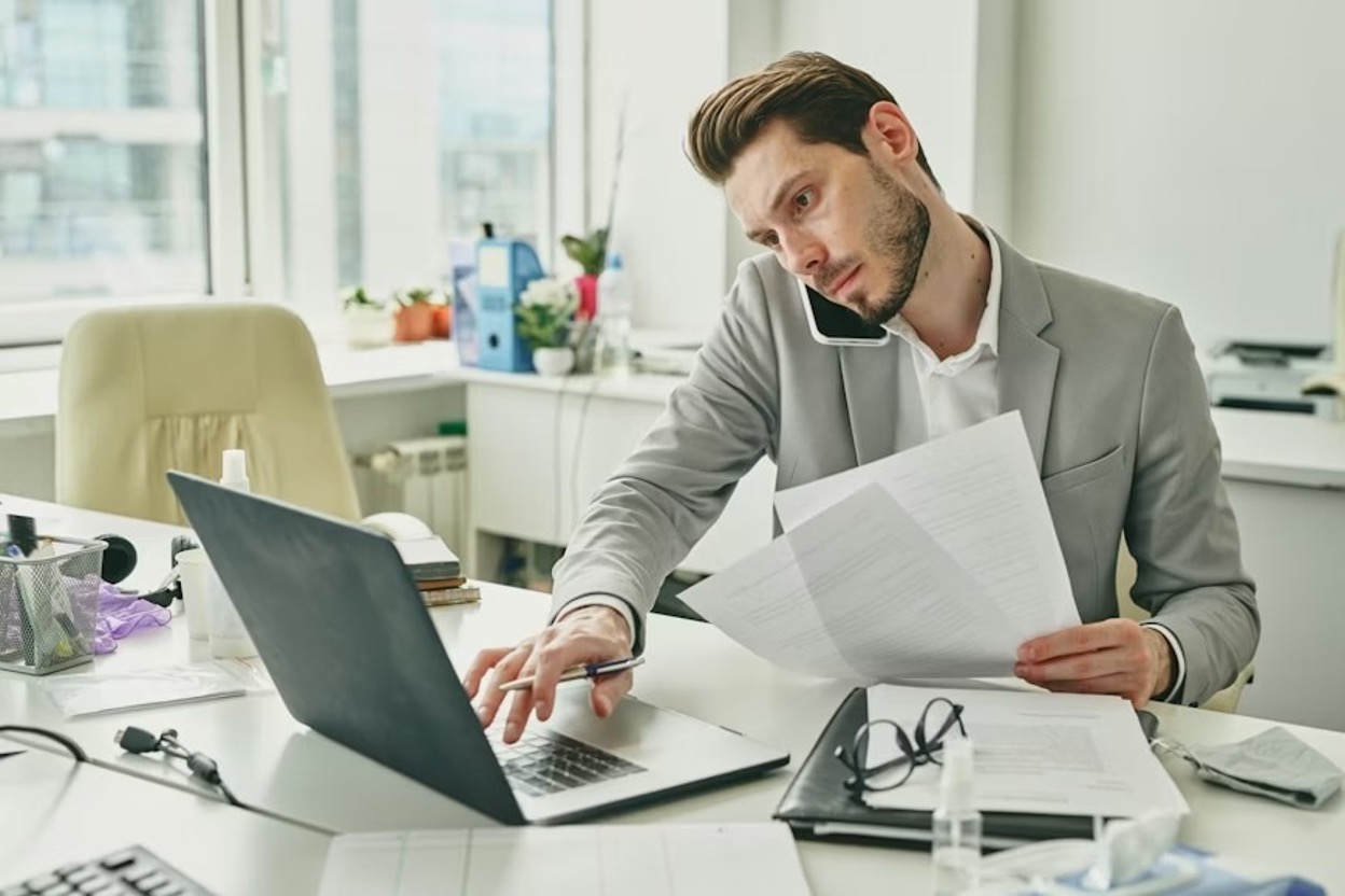 Young male office worker in formalwear consulting a client on a mobile phone while looking at content on a laptop display and going through a patent application