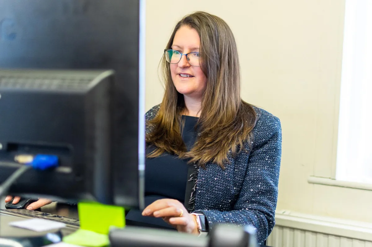 Female Mortgage Advisor with long brown hair and glasses smiling while working at a computer in an office setting.