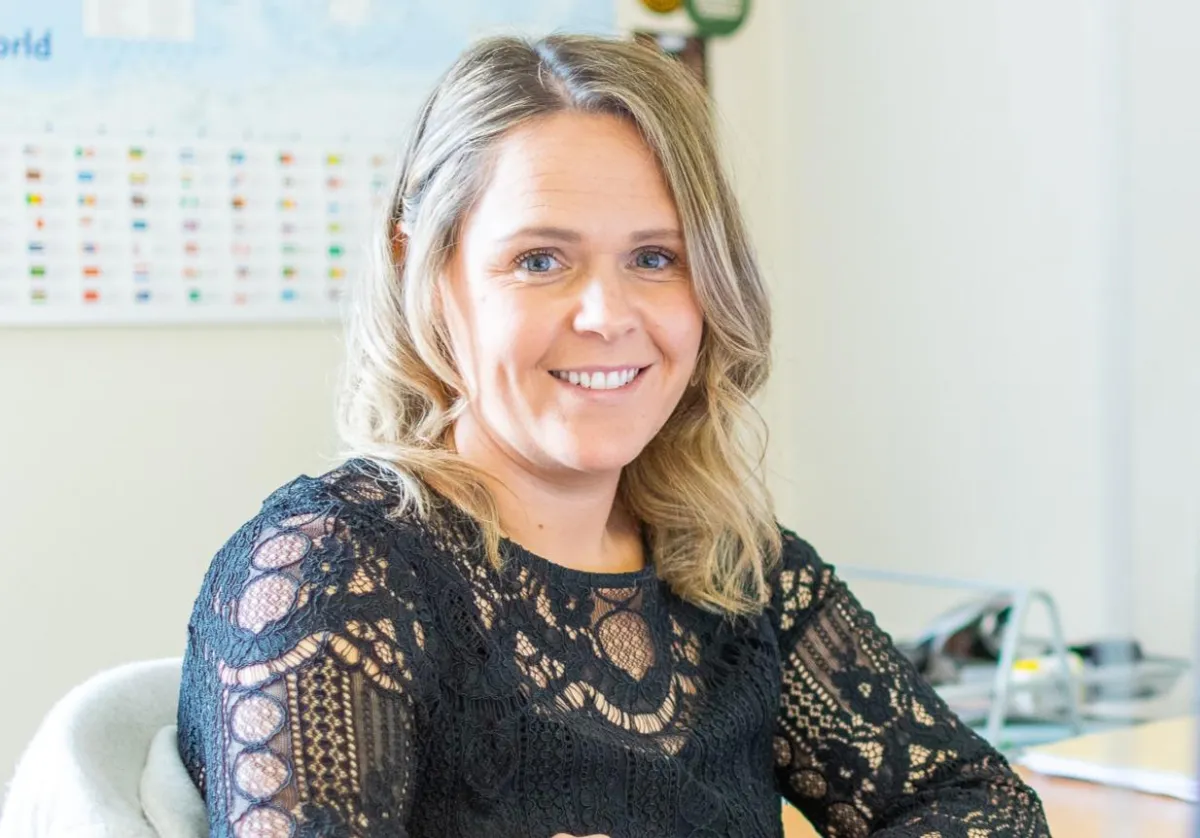 Smiling mortgage advisor with blonde wavy hair wearing a black lace top, seated at a desk in a well-lit office