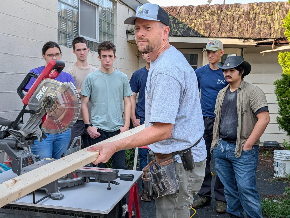 Instructor demonstrates how to cut lumber with a miter saw while Worldview Institute students watch during a hands-on Construction 101 workshop.