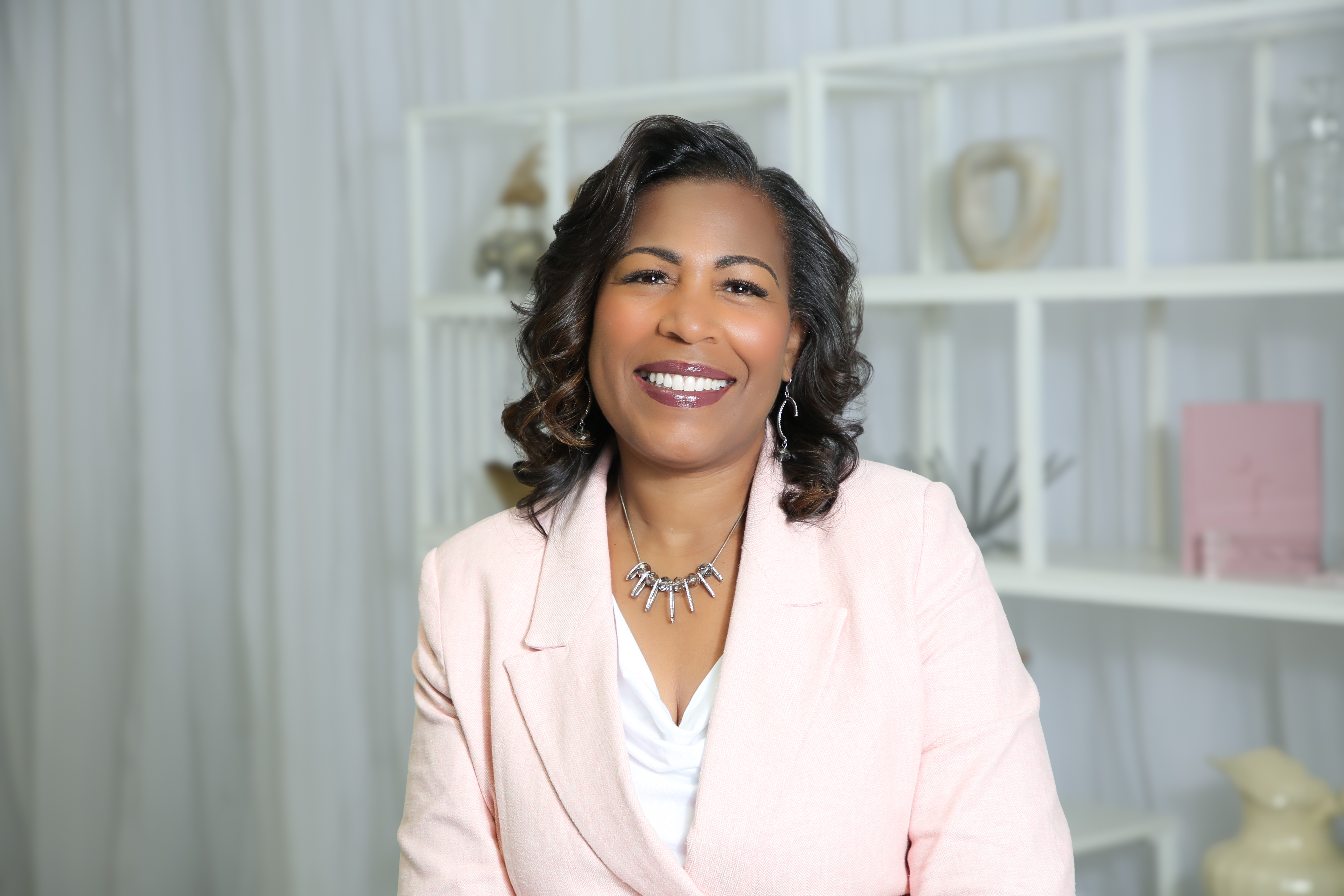 A smiling woman in her 40s with curly hair, wearing a soft pink blouse, sits in a sunlit room with books and a journal, exuding warmth and wisdom, with gentle natural light and a peaceful atmosphere.