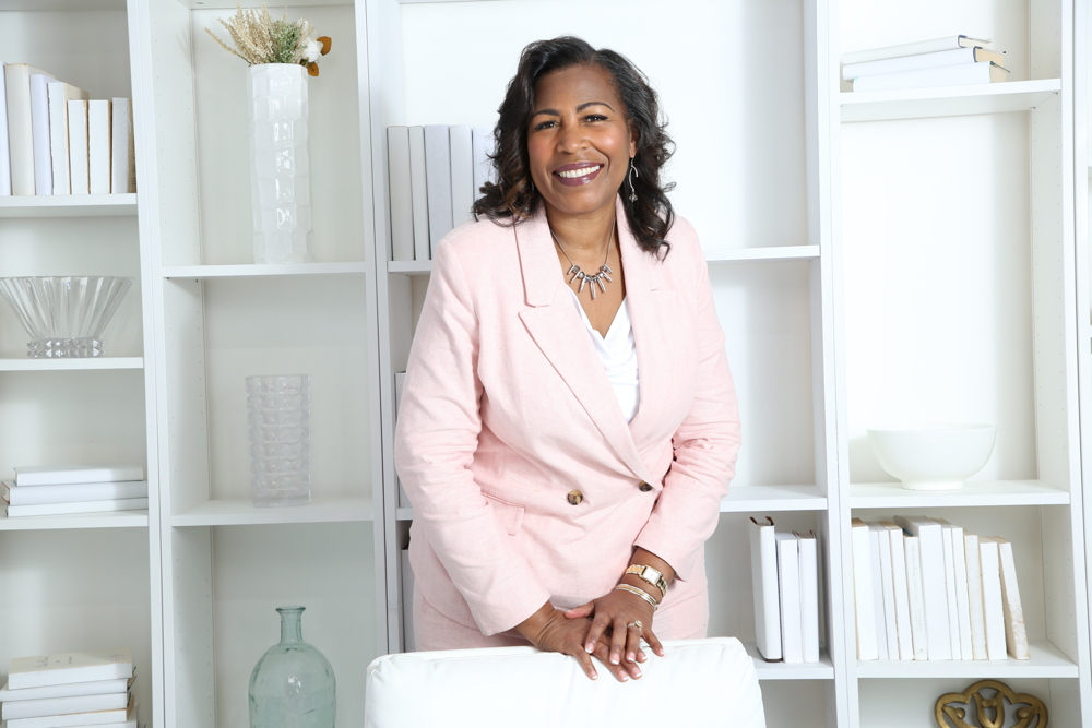 Portrait of Dr. Mona, a confident woman in her 40s with warm eyes, wearing a soft pink blouse, seated in a bright, elegant room with subtle faith-inspired decor. The setting is modern and inviting.