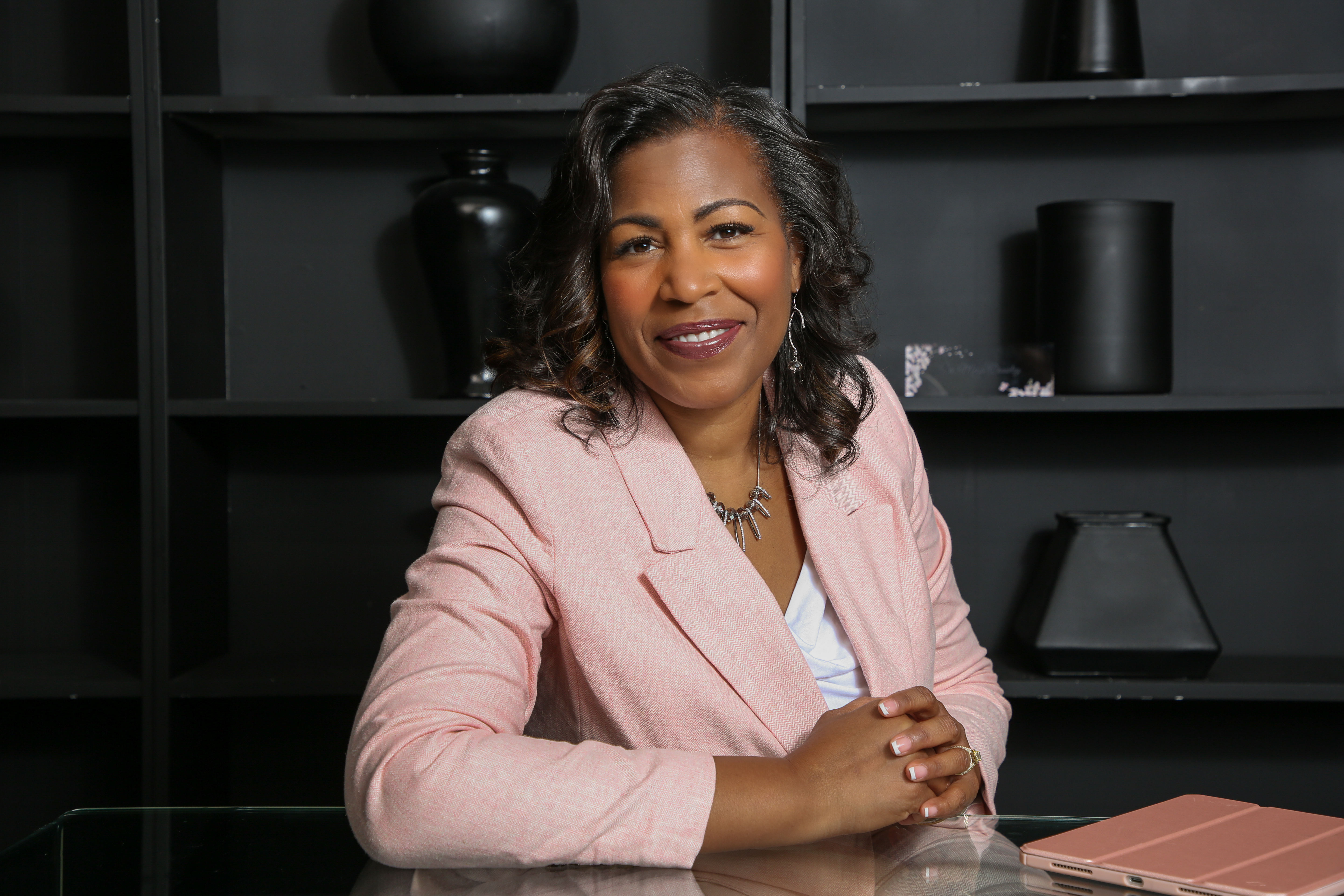 A diverse group of women in their 30s and 40s, smiling and standing together in a sunlit studio with soft pink and white decor, exuding warmth and unity.
