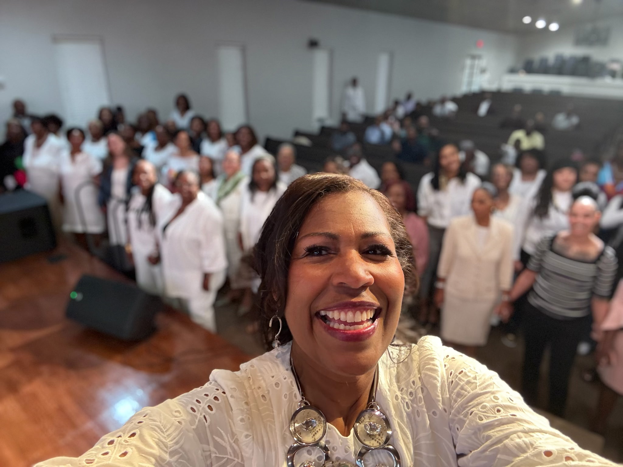 A group of five women of diverse backgrounds, seated in a circle on plush chairs in a bright, airy room with soft pink and white decor. They are laughing and sharing, creating a sense of community, support, and joyful connection.