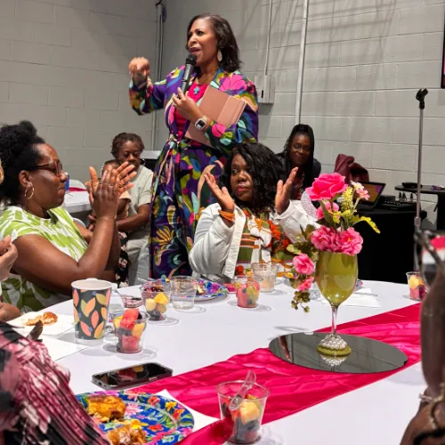 A lively workshop scene with women collaborating at a table, surrounded by notebooks, flowers, and soft pink accents in a modern space.