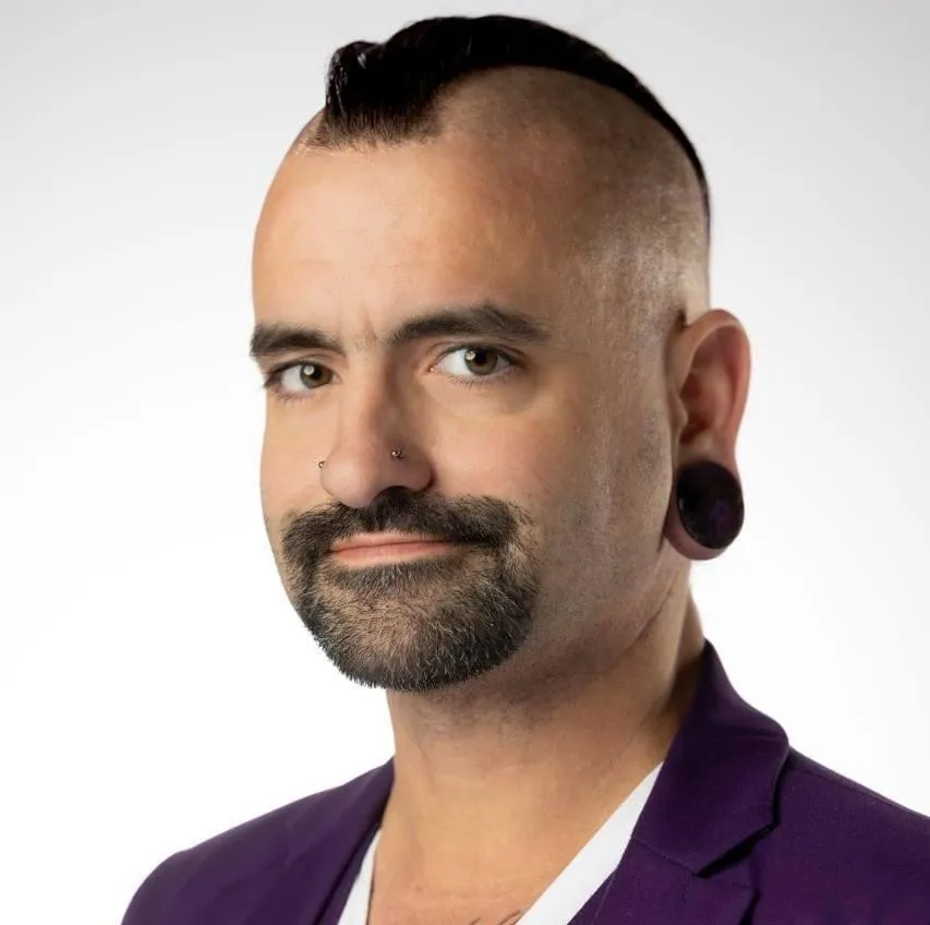 David Thorne, a white male in his 40's wearing a oatee beard. He has a mohican style haircut with shaved sides, and a goatee. He is wearing a white t-short and purple 3 piece suit. He has strecthed earlobes with dark purple plugs in