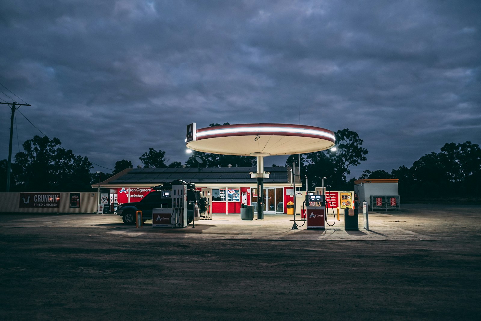 A lit truck stop and fuel station at dusk under heavy clouds, with an empty gravel lot in the foreground