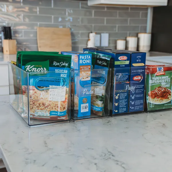 Clear kitchen organizer bins holding boxed pasta and pantry staples on a kitchen counter