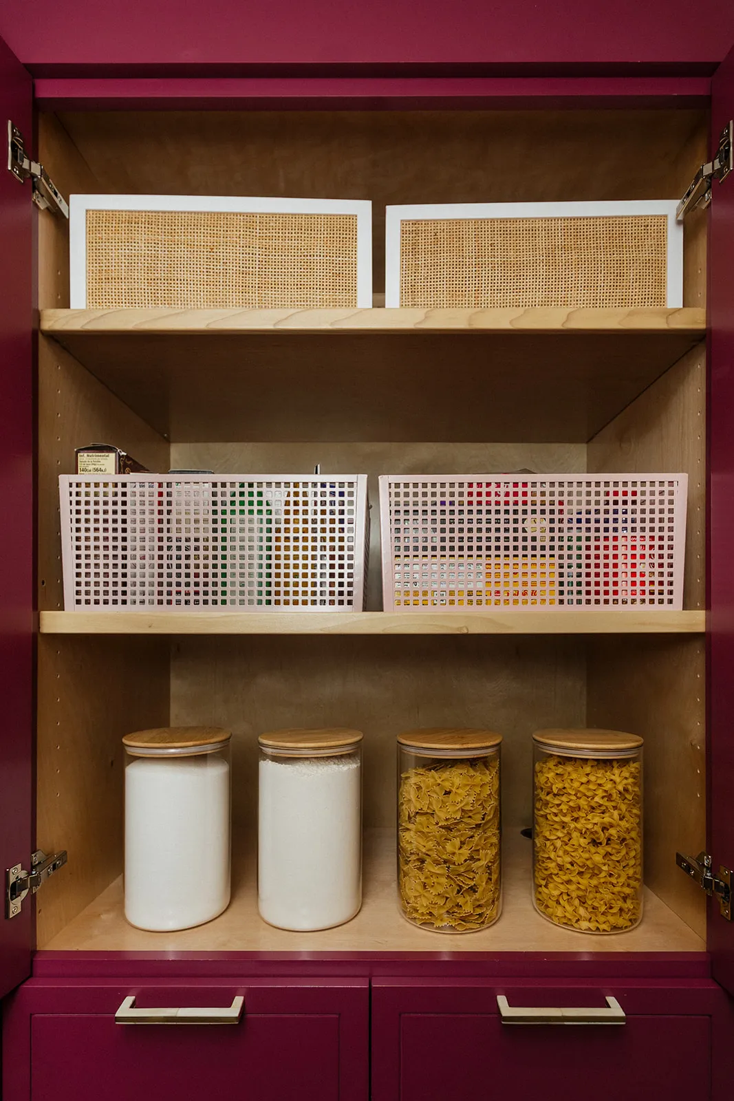 Magenta pantry cabinet organized by Tidy Crescent Co. with cane bins, pink grid baskets, and canisters of flour and pasta