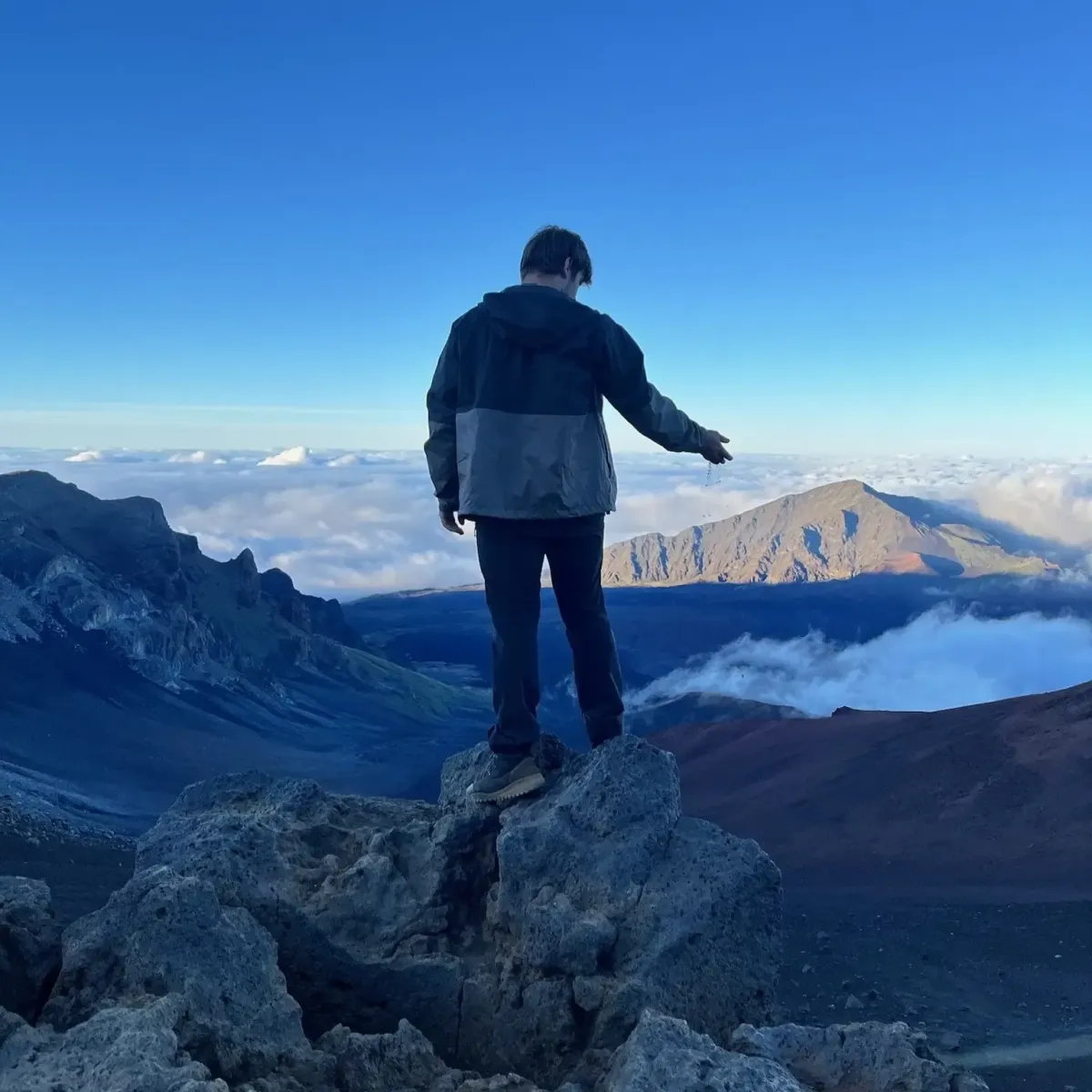 Coach Keyed standing above clouds on Maui mountain summit letting sand slip through fingers symbolizing surrender and identity level change in the 90 Day Paradigm Shift program