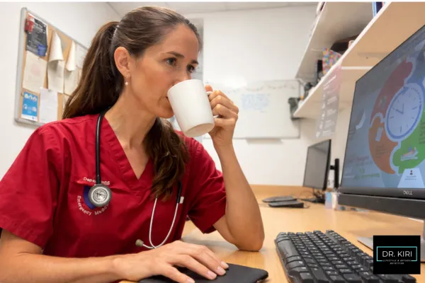 A female doctor drinking coffee while working on a computer, reflecting the busy, caffeine-fuelled reality of healthcare professionals balancing long shifts and mental fatigue.