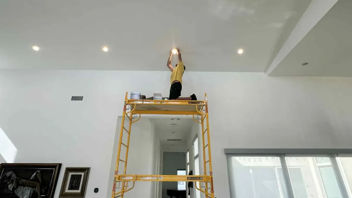 Technician on scaffolding installing recessed ceiling light in modern home interior.