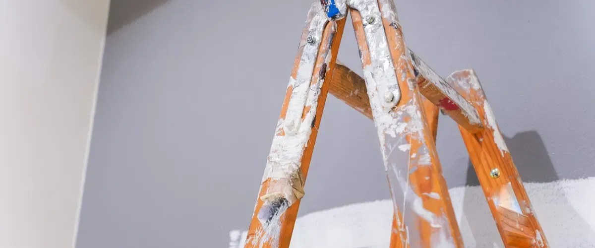 Paint-covered wooden ladder positioned against a partially painted gray wall.