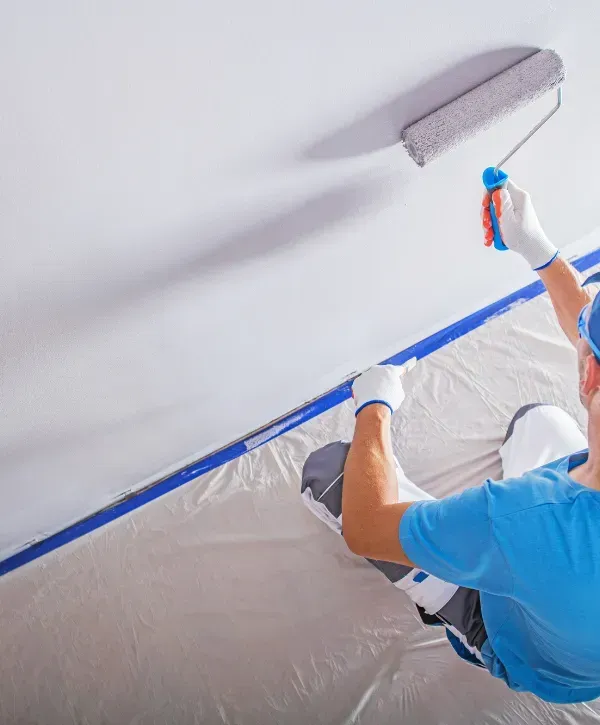 Professional painter using a roller to apply white paint to a wall while kneeling on a protected floor.