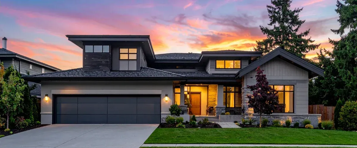 Modern suburban house exterior at sunset with warm interior lighting and landscaped yard.