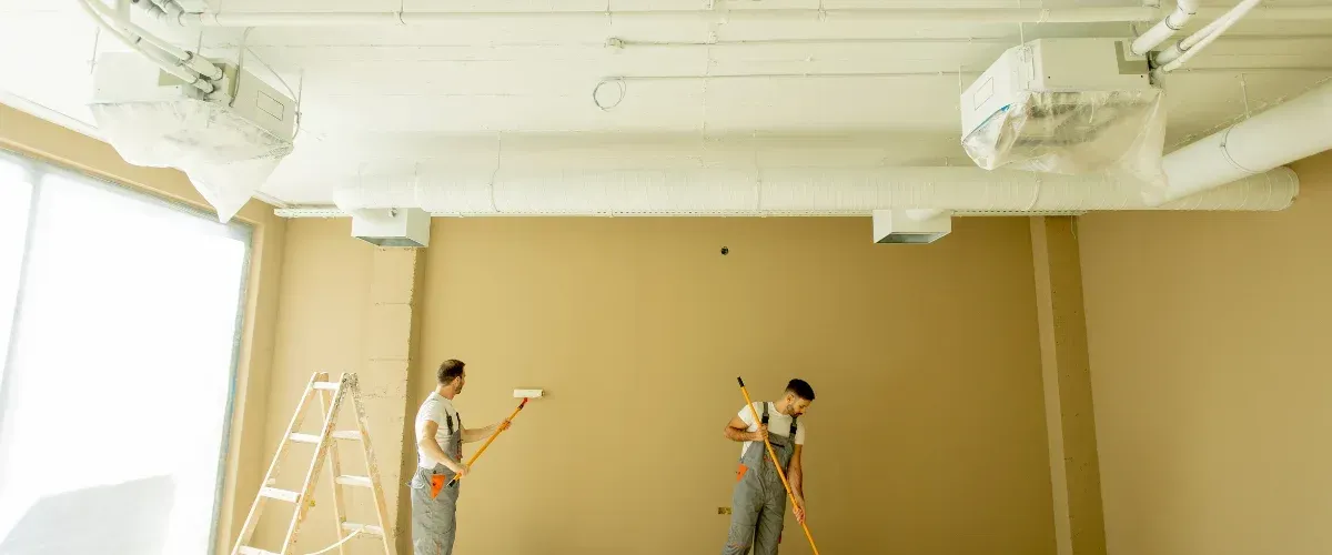 Two professional painters applying beige paint to an interior wall in a commercial space.