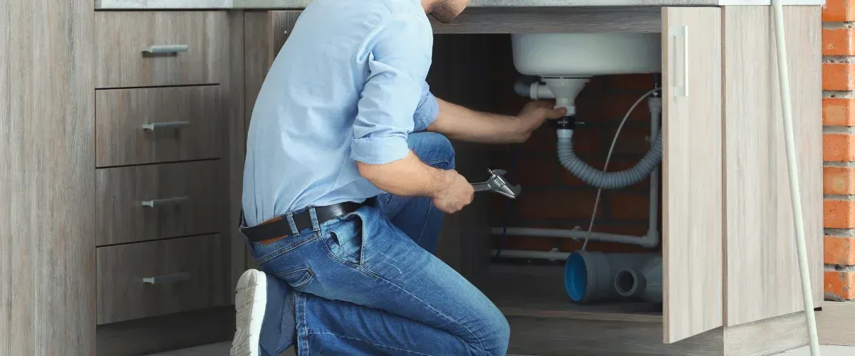 A man in a blue shirt and jeans kneels, using a wrench to fix the plumbing pipes under a kitchen sink.