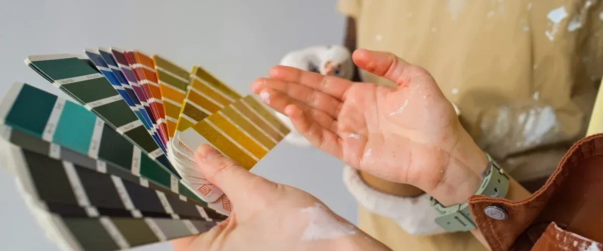 A close-up of two hands, one holding a fan of paint swatches and the other gestured mid-air, both spattered with paint.