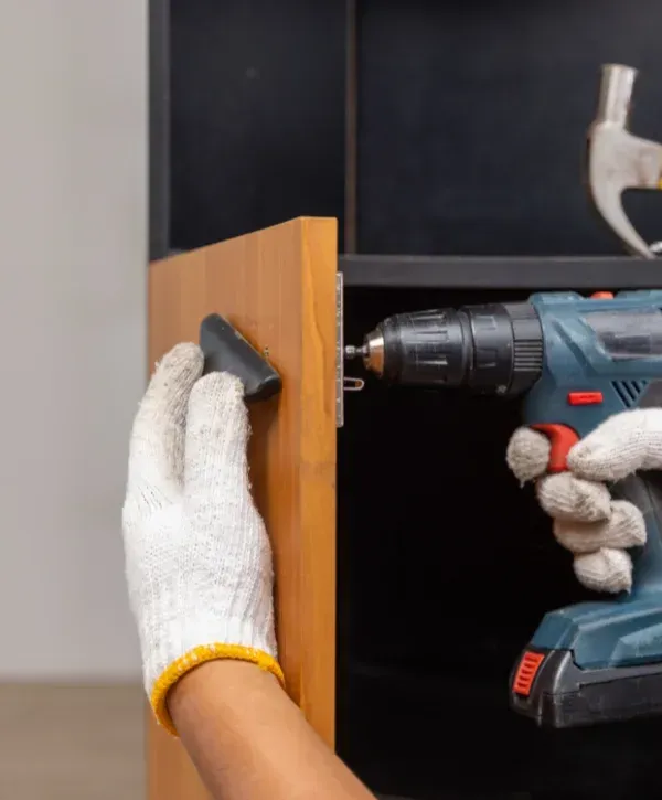 A gloved hand holds a black cabinet handle while another gloved hand uses a drill to install it.
