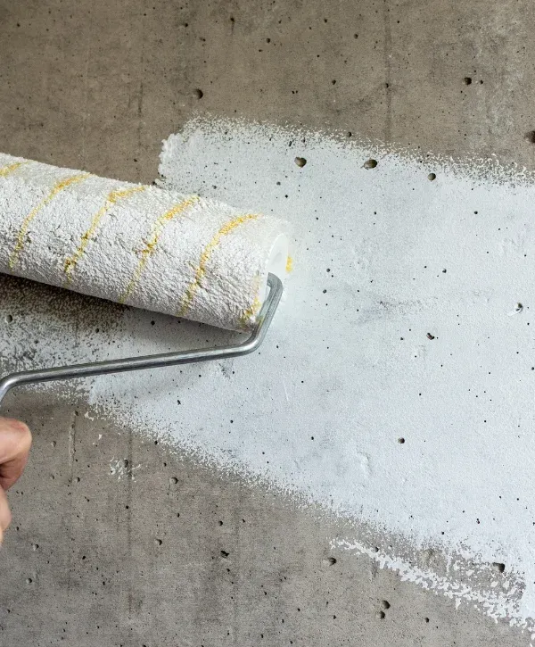A paint roller applying white primer to a rough, grey concrete wall, with a hand holding the roller.