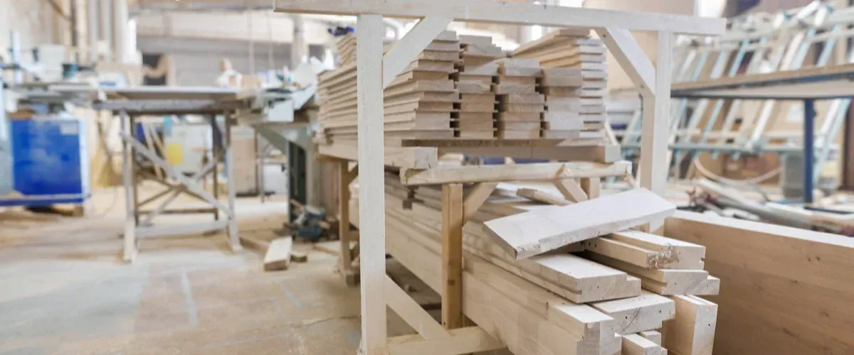 Stacks of light-colored sawn timber and wood planks on wooden racks in a blurred carpentry workshop.
