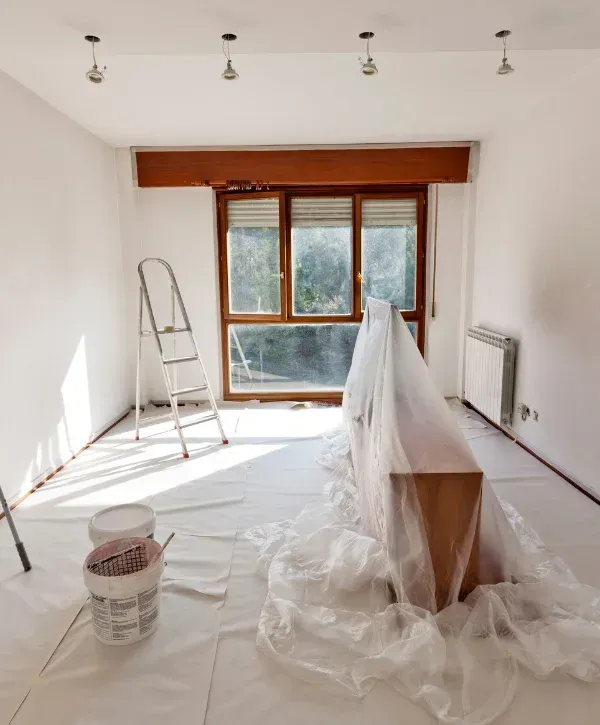 An empty room prepared for renovation with a ladder, paint buckets, and covered furniture near a large window.