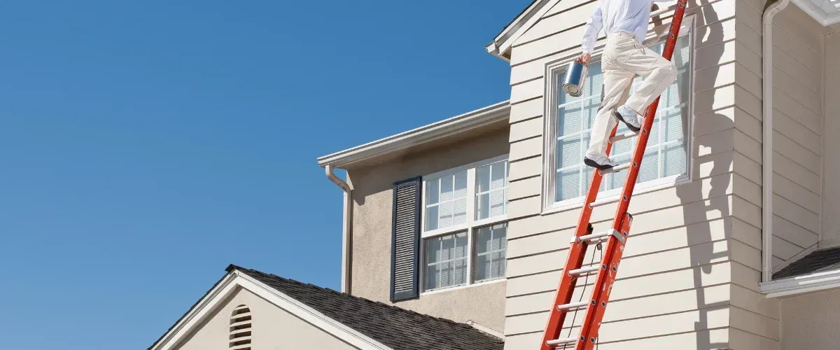 A person in khaki pants and a white shirt ascends an orange ladder, holding a paint can, to paint a cream-colored house.