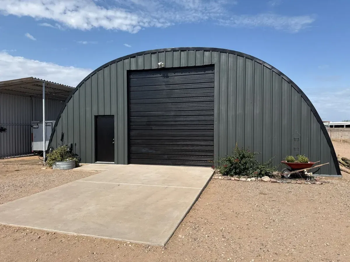 A dark green Quonset hut with a black rolling door and small black entry door, under a blue sky.