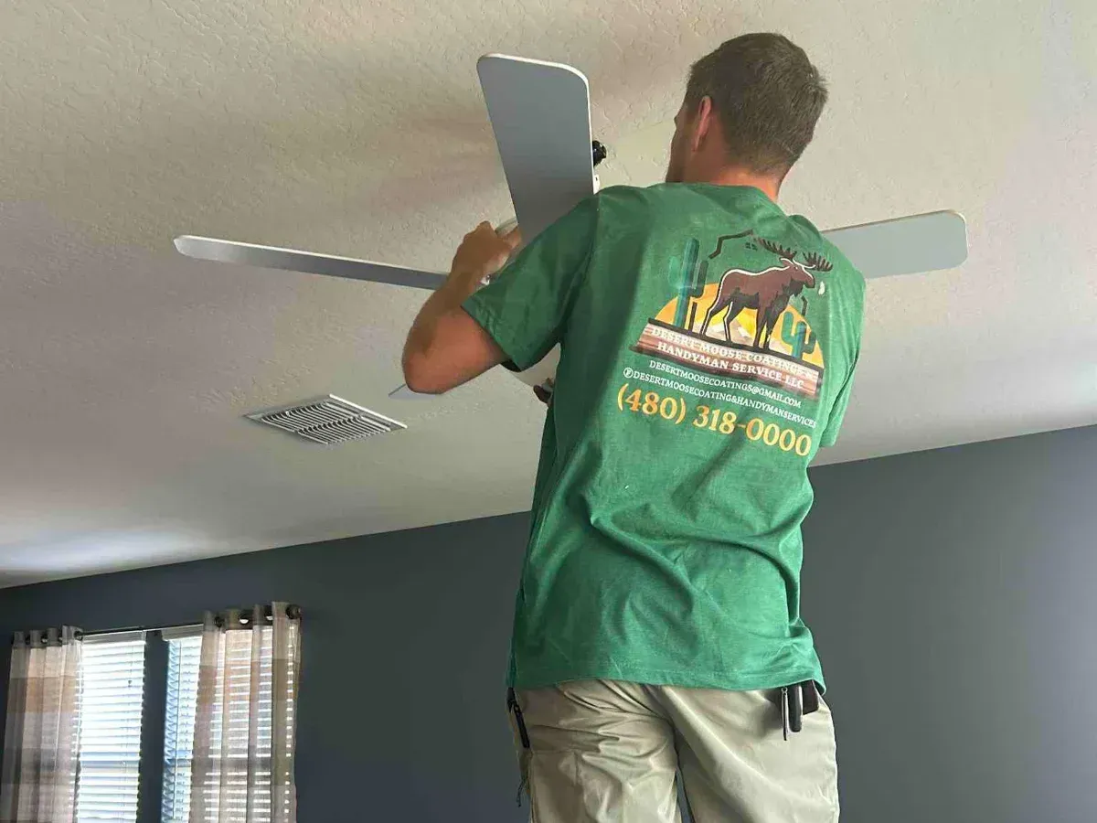 A handyman in a green t-shirt installs a white ceiling fan, seen from behind his shoulder, fixing the blade.