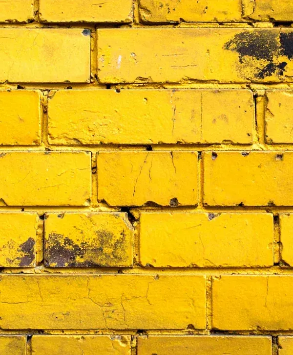 A close-up view of a distressed brick wall painted a vibrant yellow, with black marks and cracked paint.